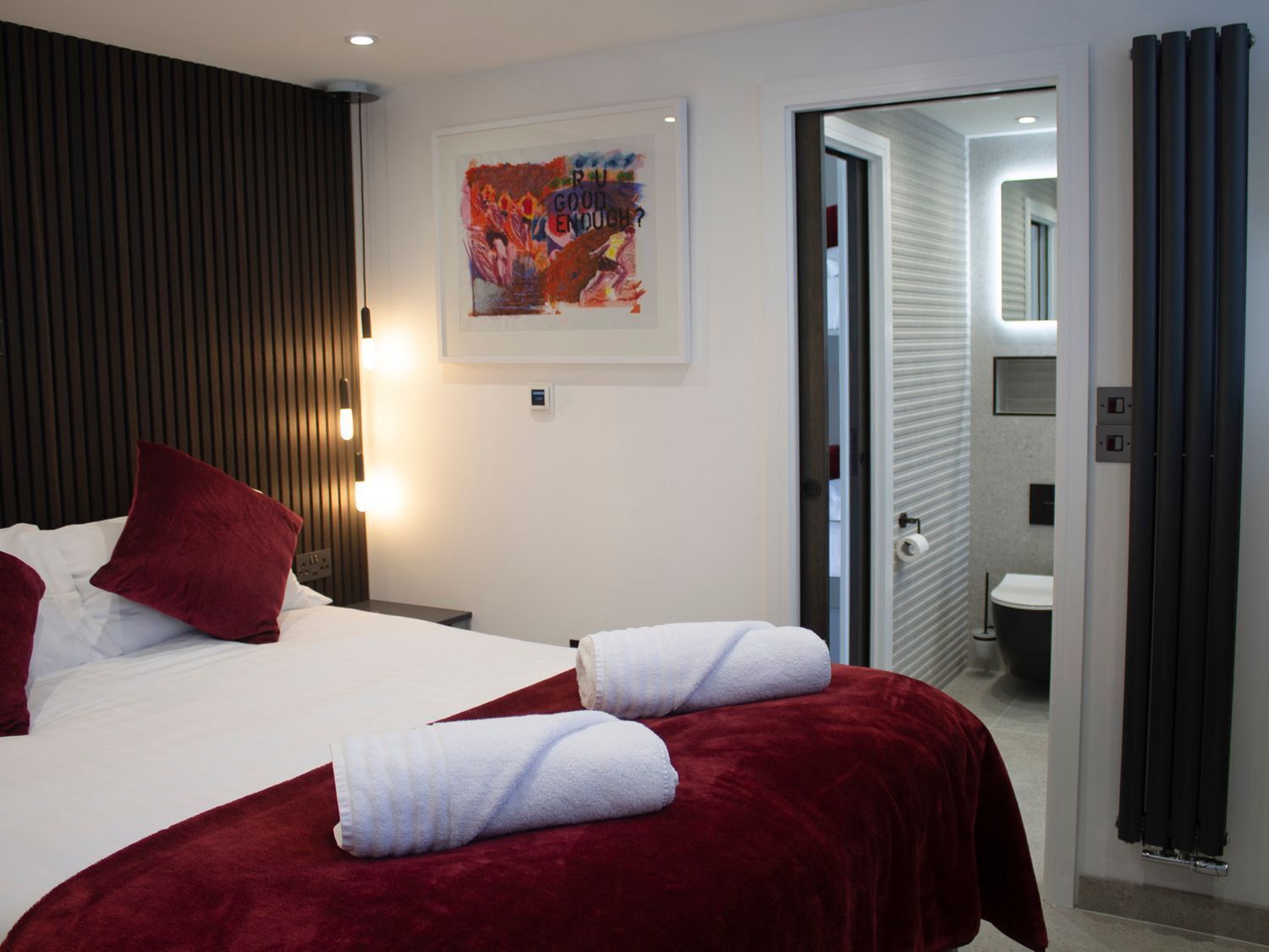 Habitación de hotel con una cama, dos almohadas rojas, una colcha blanca y un plaid rojo. Se ve un cuadro en la pared y un baño a través de una puerta abierta.