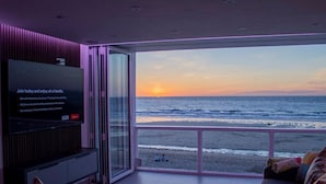 Un salon moderne avec un grand écran de télévision affichant le logo de Netflix, une porte vitrée donnant sur un balcon avec vue sur l’océan au coucher du soleil.