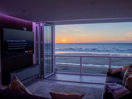 Un salón moderno con un televisor grande mostrando el logotipo de Netflix, una puerta de vidrio que da a un balcón con vista al océano al atardecer.