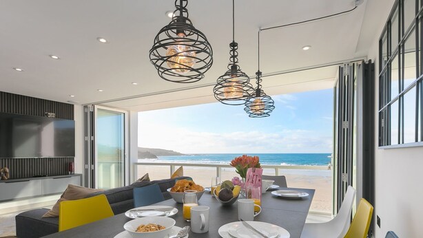 Un espace repas moderne avec vue sur la plage, comprenant une longue table dressée avec des assiettes, des bols et une carafe de jus d’orange.