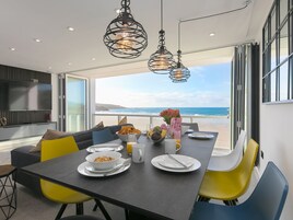 Un moderno comedor con vista a la playa, que presenta una mesa larga con platos, tazones y una jarra de jugo de naranja.