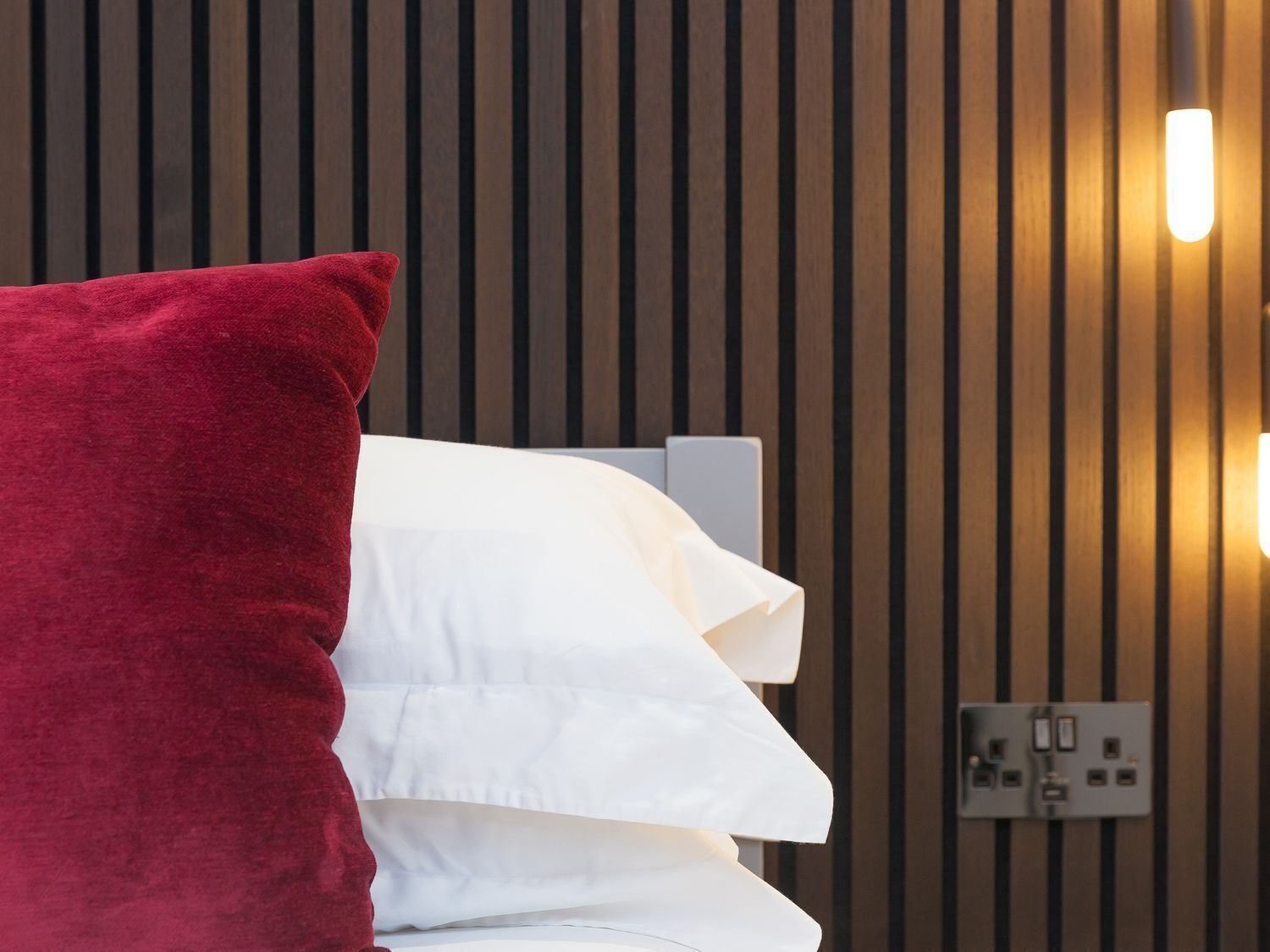 A bed with white and red pillows against a wooden slat wall with a wall socket.