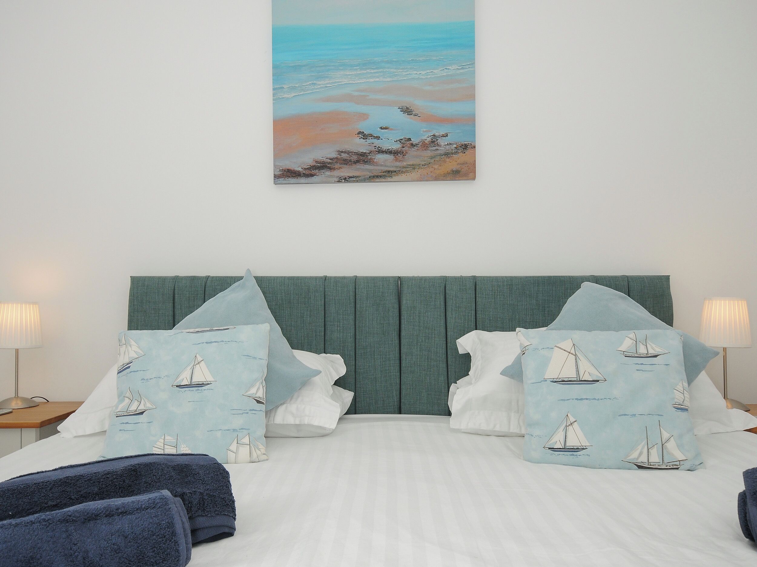 A hotel room with a bed, two nightstands, a painting of a beach scene, and pillows with sailboat designs.
