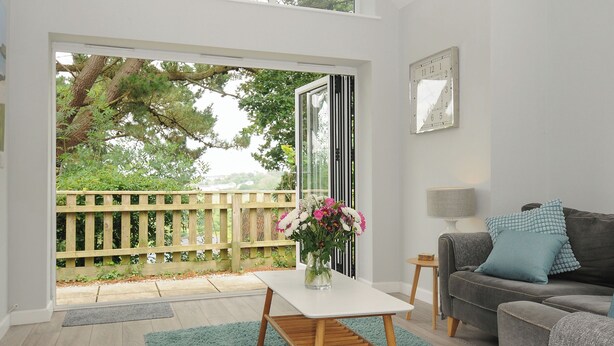 A modern living room with a white table, a grey sofa, and a view of a wooden deck and greenery outside.