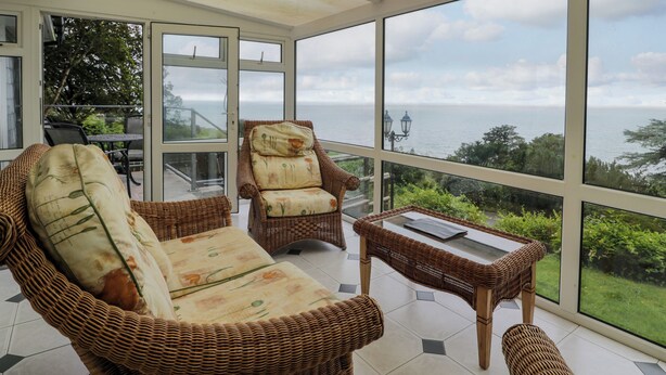 A sunlit room with wicker furniture, large windows, and a view of the sea. - Woodland Roost (Barmouth)