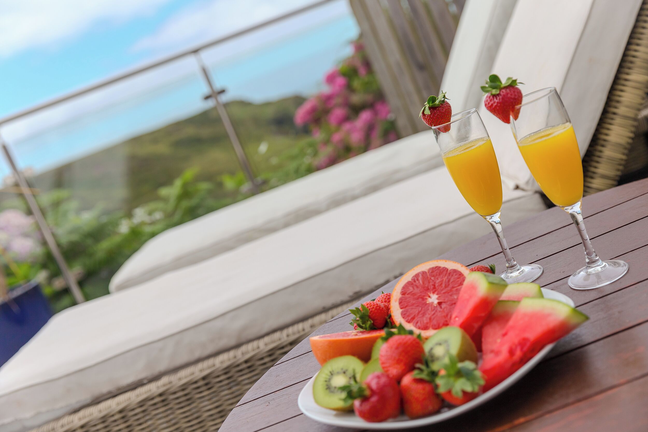 A table with a fruit platter and two glasses of orange juice, set on a balcony with a view of the sea and greenery.