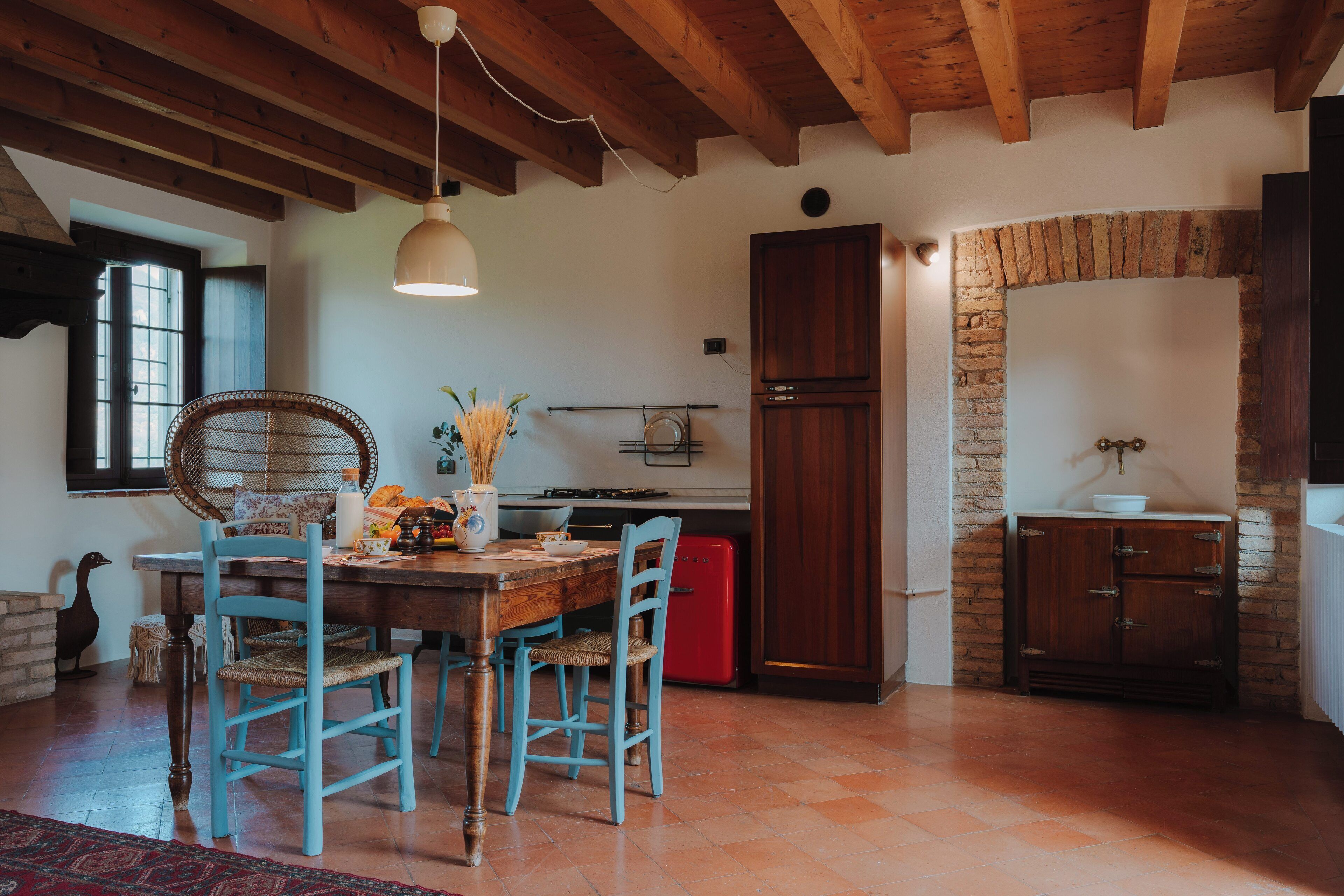 Une cuisine rustique avec des poutres en bois, une table à manger entourée de chaises bleues, un réfrigérateur rouge et un mur en pierre.
