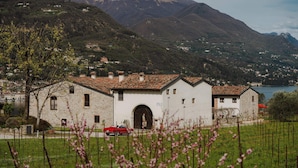 A stone building with a red car parked in front, surrounded by greenery and mountains in the background. - L'Angelica Corte Palazzina (Salò)