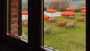 A view through a window with a brick wall, a pool area with red umbrellas, and mountains in the background. - L'Angelica Corte Palazzina (Salò)