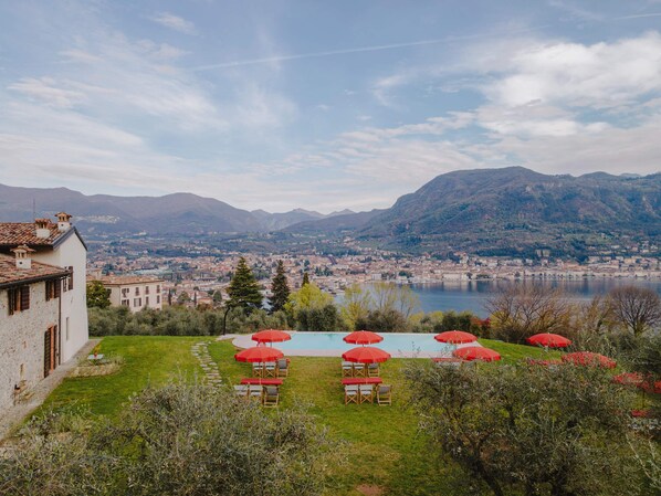 Zona de piscina con sombrillas rojas y sillas de descanso, rodeada de vegetación y con vistas a un paisaje urbano y montañas al fondo.