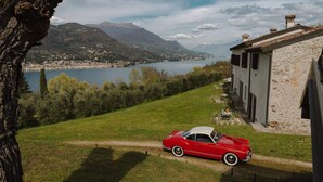 A red vintage car parked on a grassy area with a stone building and a lake in the background. - L'Angelica Corte Palazzina (Salò)