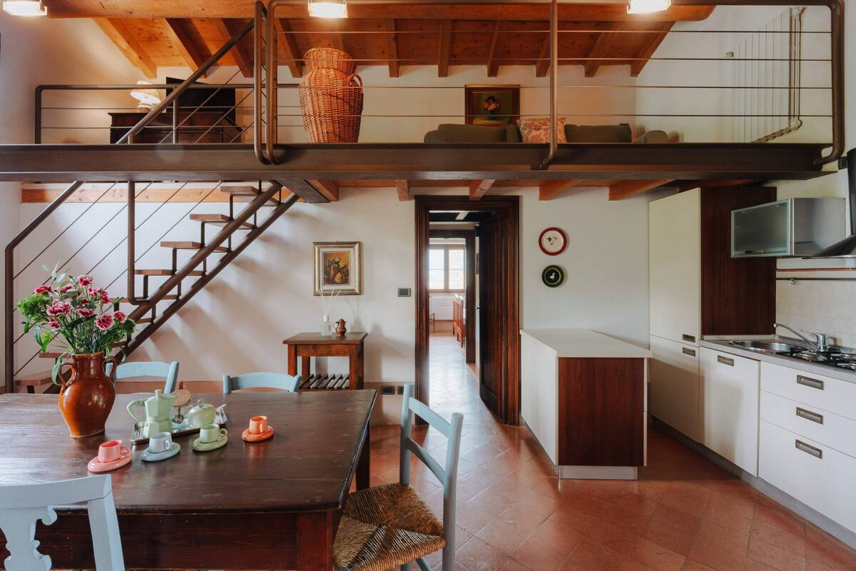 Un'ampia cucina con soffitto alto, un'area pranzo con tavolo e sedie in legno, e una scala che collega due livelli.