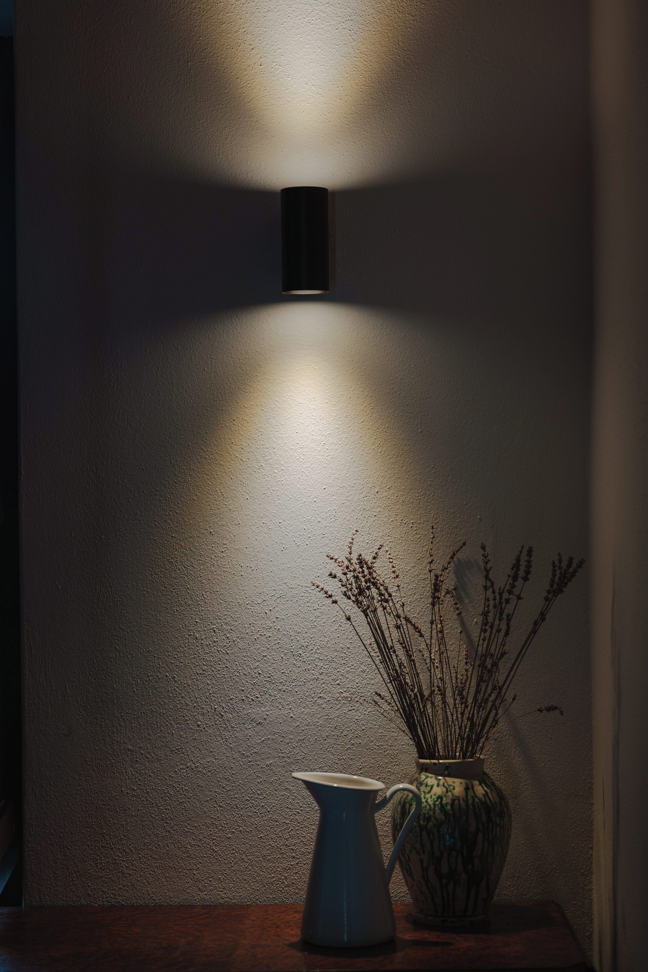 A wall-mounted light fixture casting a shadow on a textured wall, a ceramic vase with dried plants, and a white pitcher on a wooden surface.