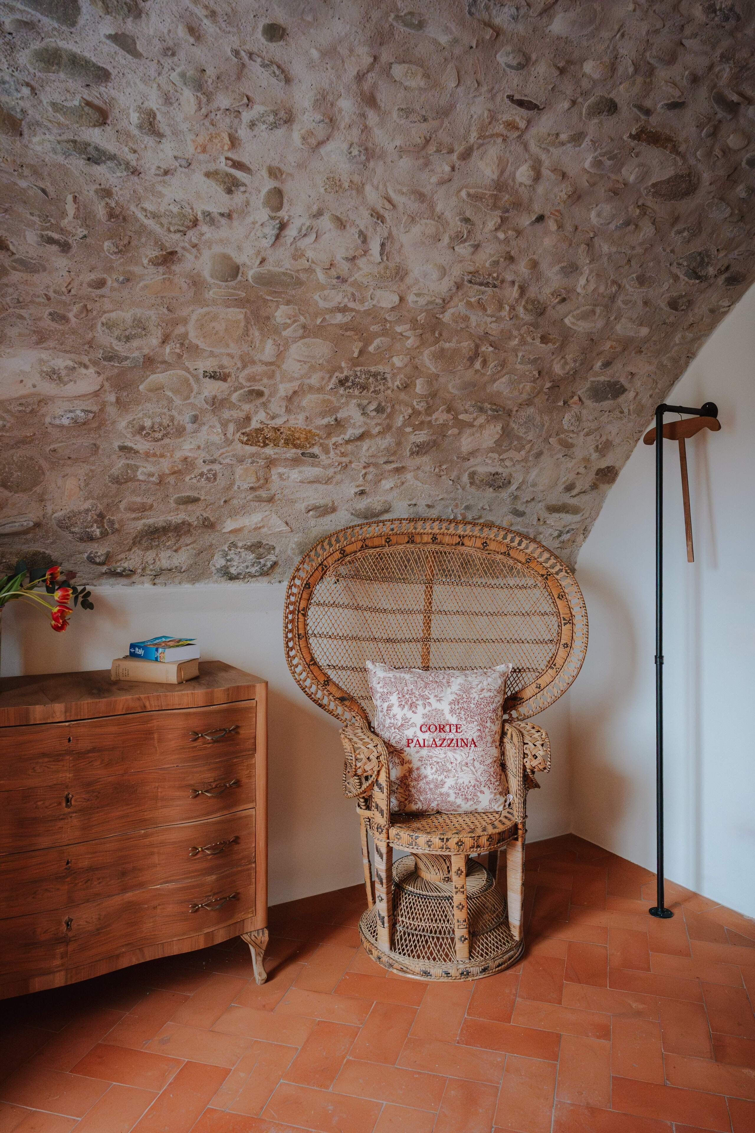 A rustic room with a stone ceiling, a wicker chair with a cushion, a wooden dresser, and a floor lamp.