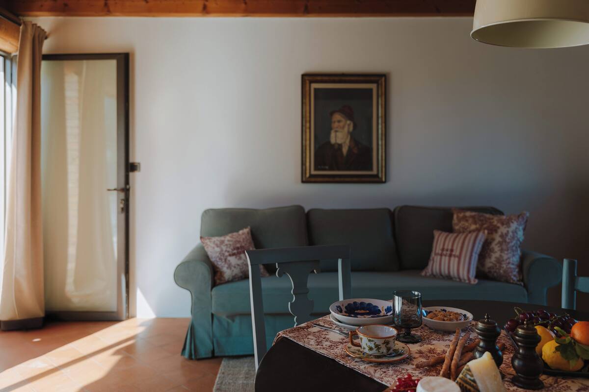 Un soggiorno accogliente con un divano color petrolio, un tavolo da pranzo con tovaglia floreale e un quadro appeso al muro.
