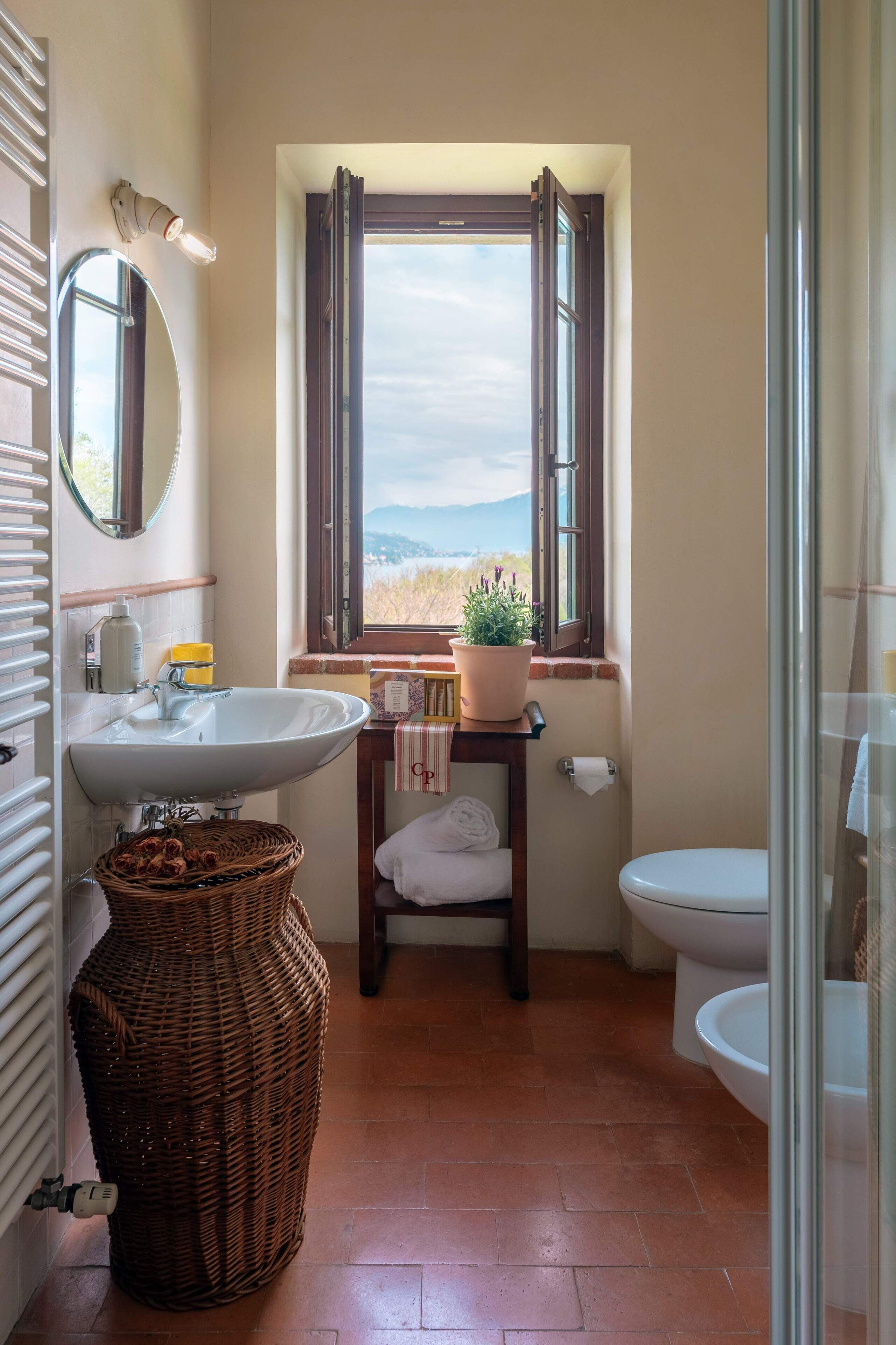 A bathroom with a large sink, a mirror, a window with a view, a toilet, and a wicker basket.