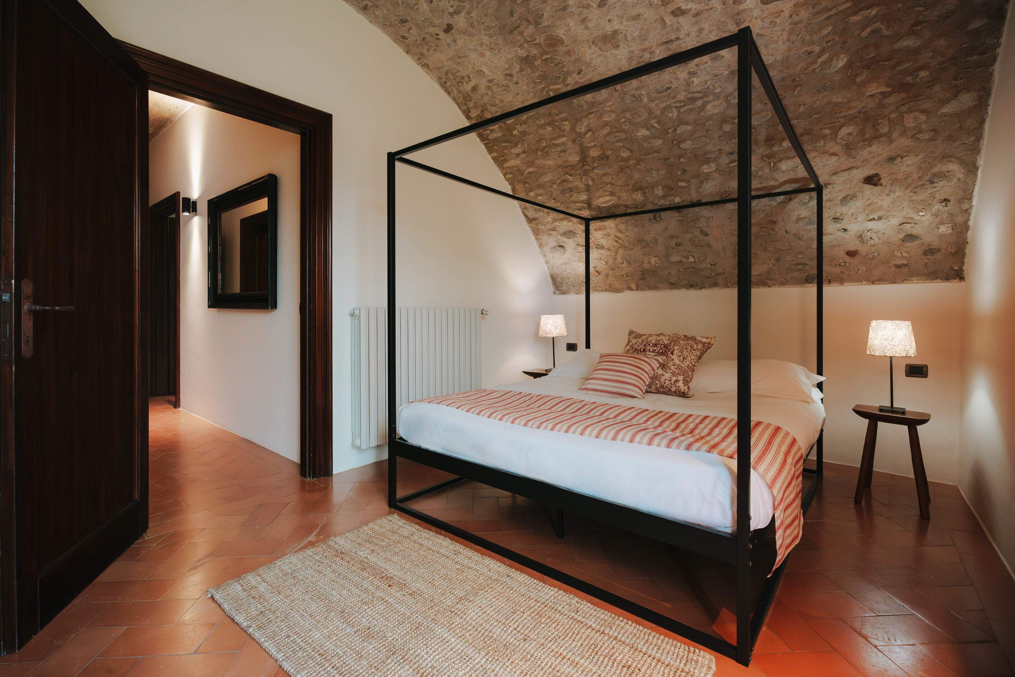 Una cama con dosel en una habitación con techo de piedra, suelo de madera y una mesita de noche con lámpara.