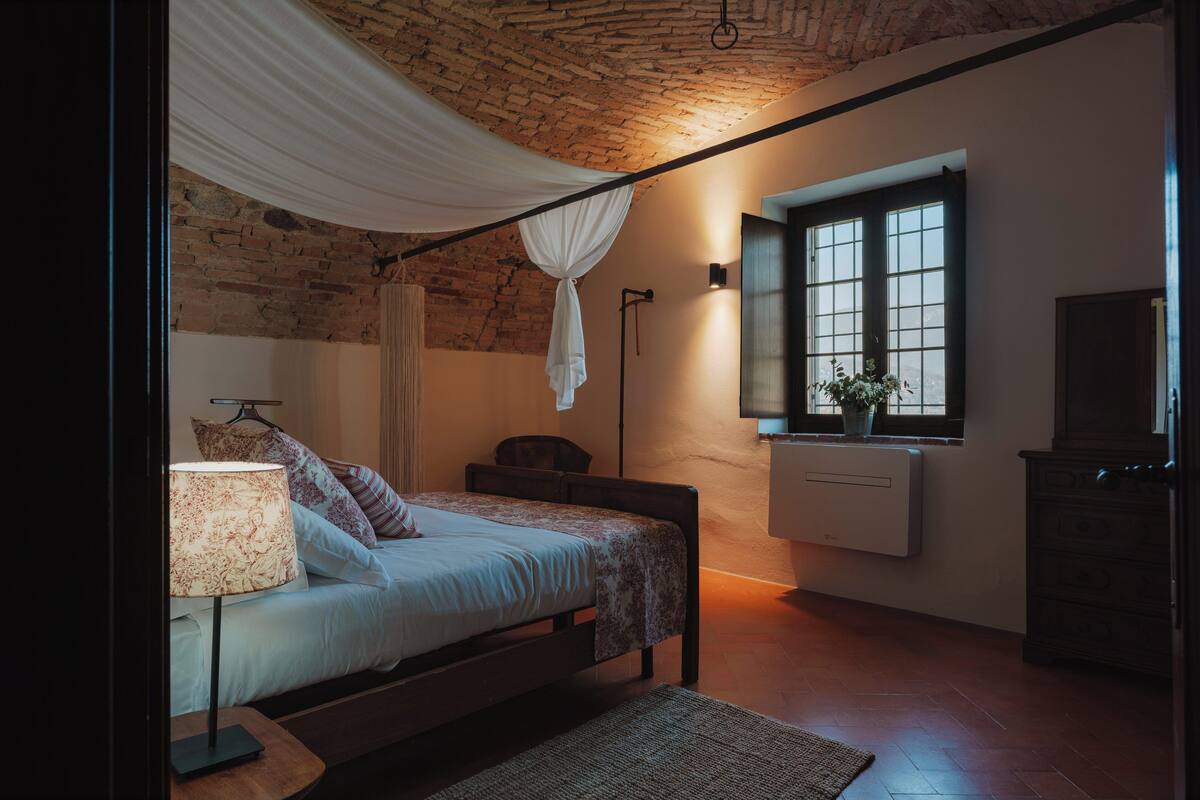 Una camera da letto con soffitto in mattoni, un letto con baldacchino bianco, una lampada da comodino, un termosifone, una cassettiera e una finestra con vista.