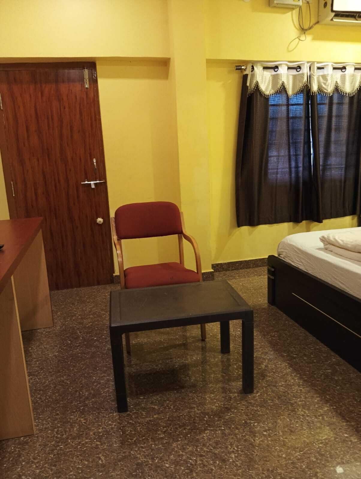 Een kleine kamer met een houten deur, een rode stoel, een klein tafeltje, een bed en een raam met gordijnen.