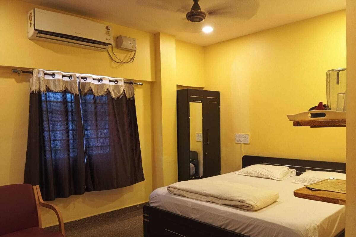 A hotel room with a bed, a chair, an air conditioning unit, a mirror, and a shelf with a glass.