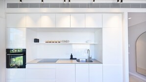 Een moderne keuken met witte kastjes, een inbouwoven, een roestvrijstalen spoelbak en een aanrecht met een koffieapparaat en kopjes.