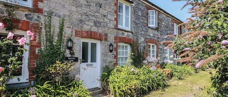 A stone cottage with a white door, a small garden with various plants, and a clear sky.