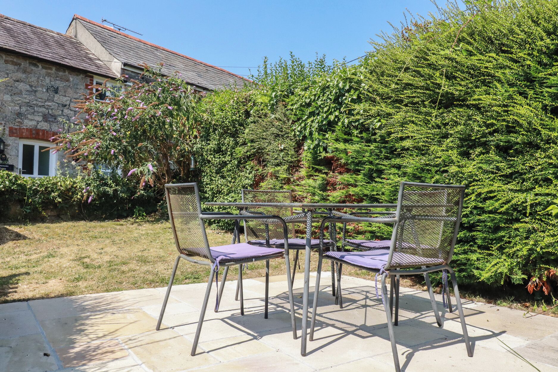 A patio with a table and four chairs, surrounded by a stone wall and a dense green hedge.
