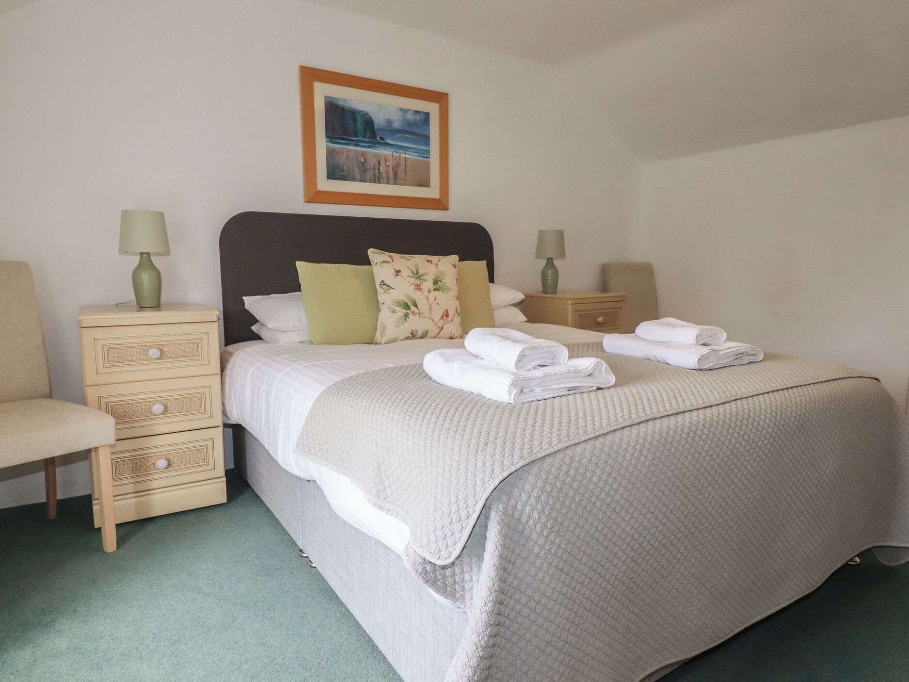 A neatly made bed with a quilted cover, two bedside tables with lamps, a chair, and a framed picture on the wall.