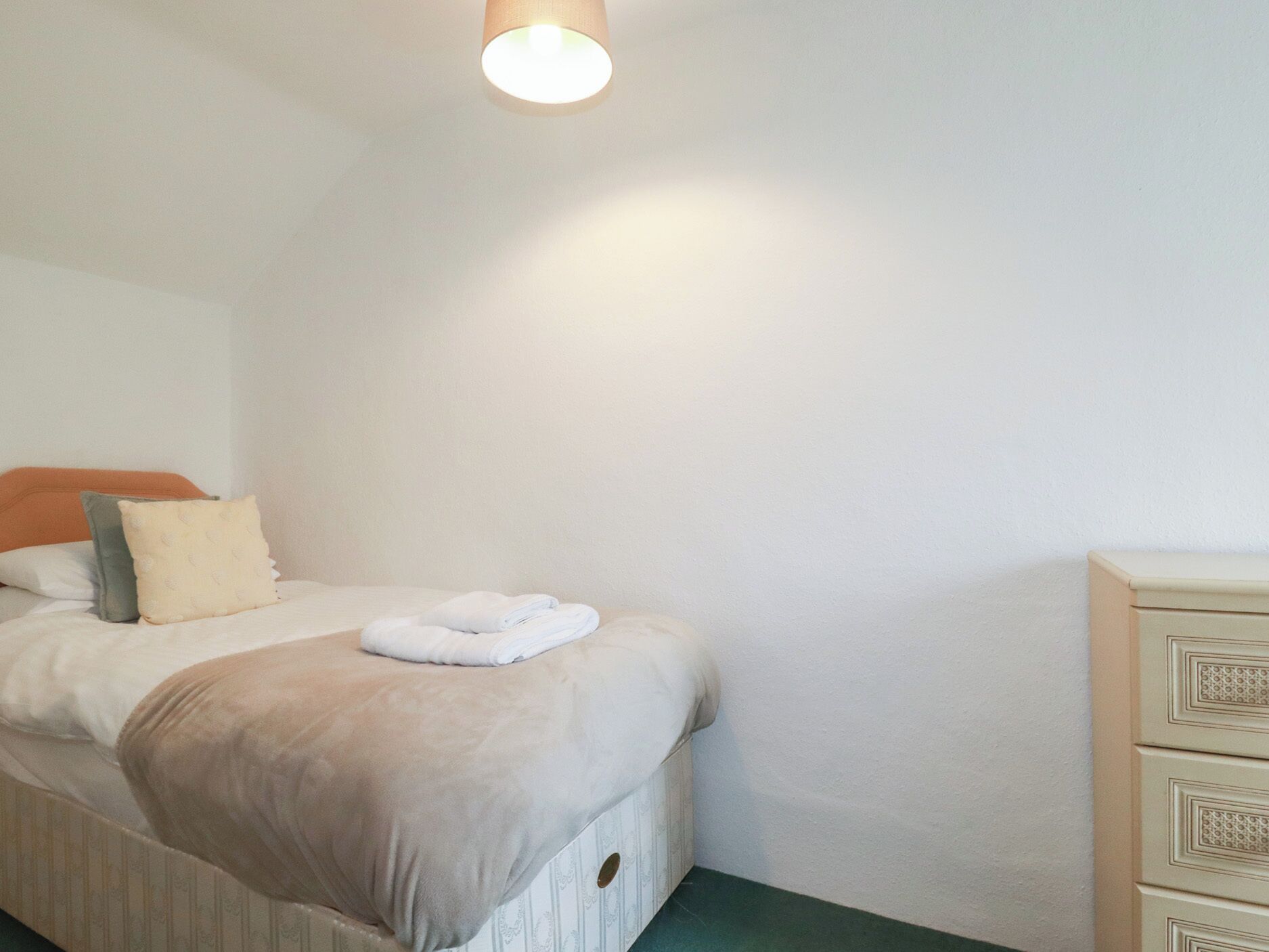 A neatly made bed with a white comforter and a folded towel, a bedside table with a drawer, and a plain wall with a ceiling light.