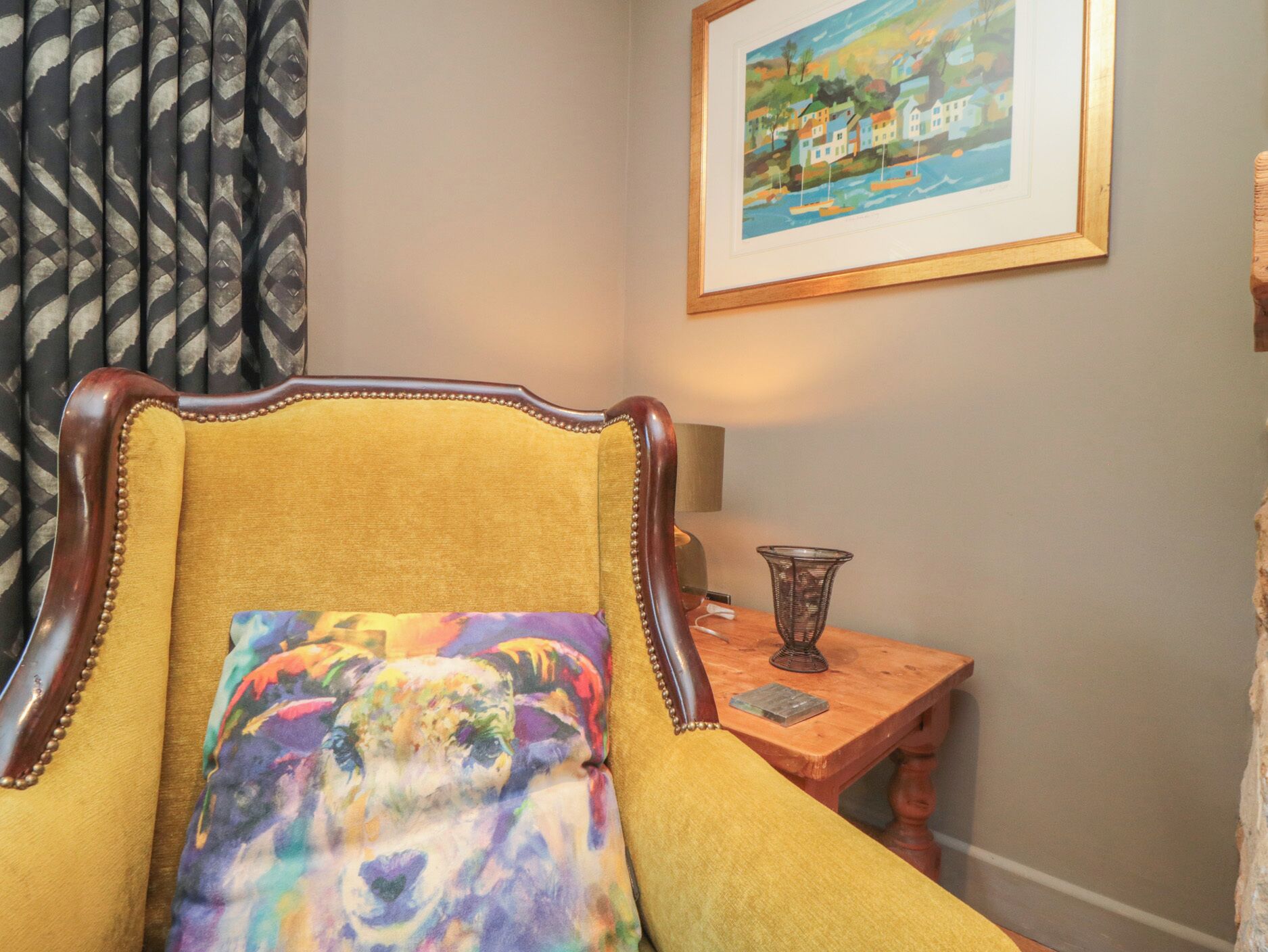 A room with a yellow armchair, a framed painting on the wall, and a small wooden table with a lamp.