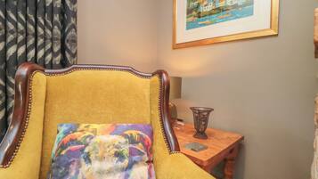 A room with a yellow armchair, a framed painting on the wall, and a small wooden table with a lamp.
