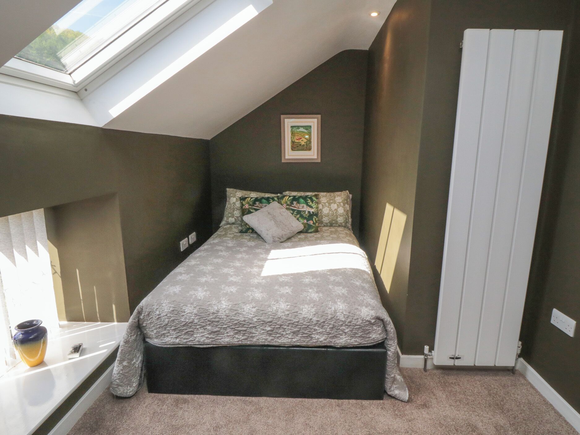A bedroom with a skylight, a bed with patterned bedding, a radiator, and a small window with a vase.