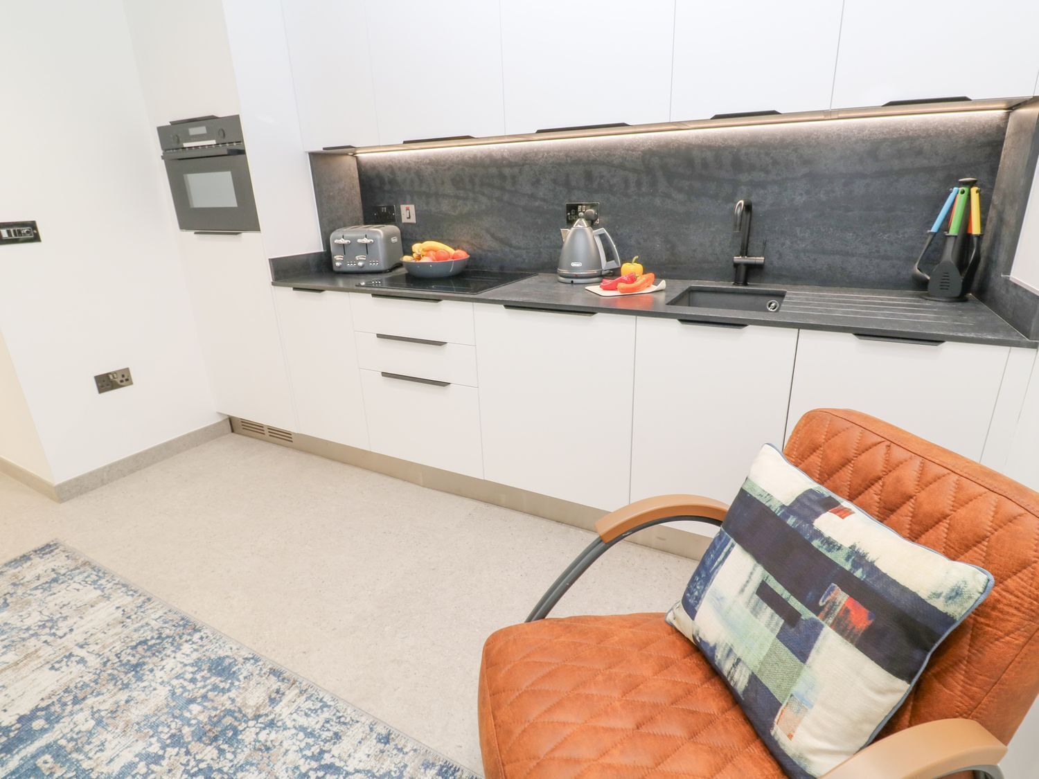 Cocina compacta con estufa integrada, un lavamanos pequeño y una encimera con tetera y tostadora. Hay una silla con un cojín acolchado y un almohadón estampado.