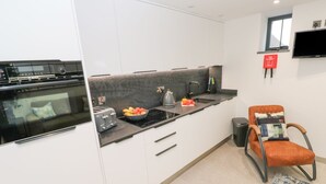 Una cocina moderna con horno empotrado, encimera negra y un bol de fruta. Se observa un cartel de manta ignífuga roja en la pared y una silla con cojín.
