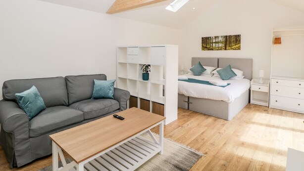 Un dormitorio moderno con un sofá gris, una mesa de centro de madera, un armario blanco, una cama con ropa de cama azul y un cuadro de un bosque.