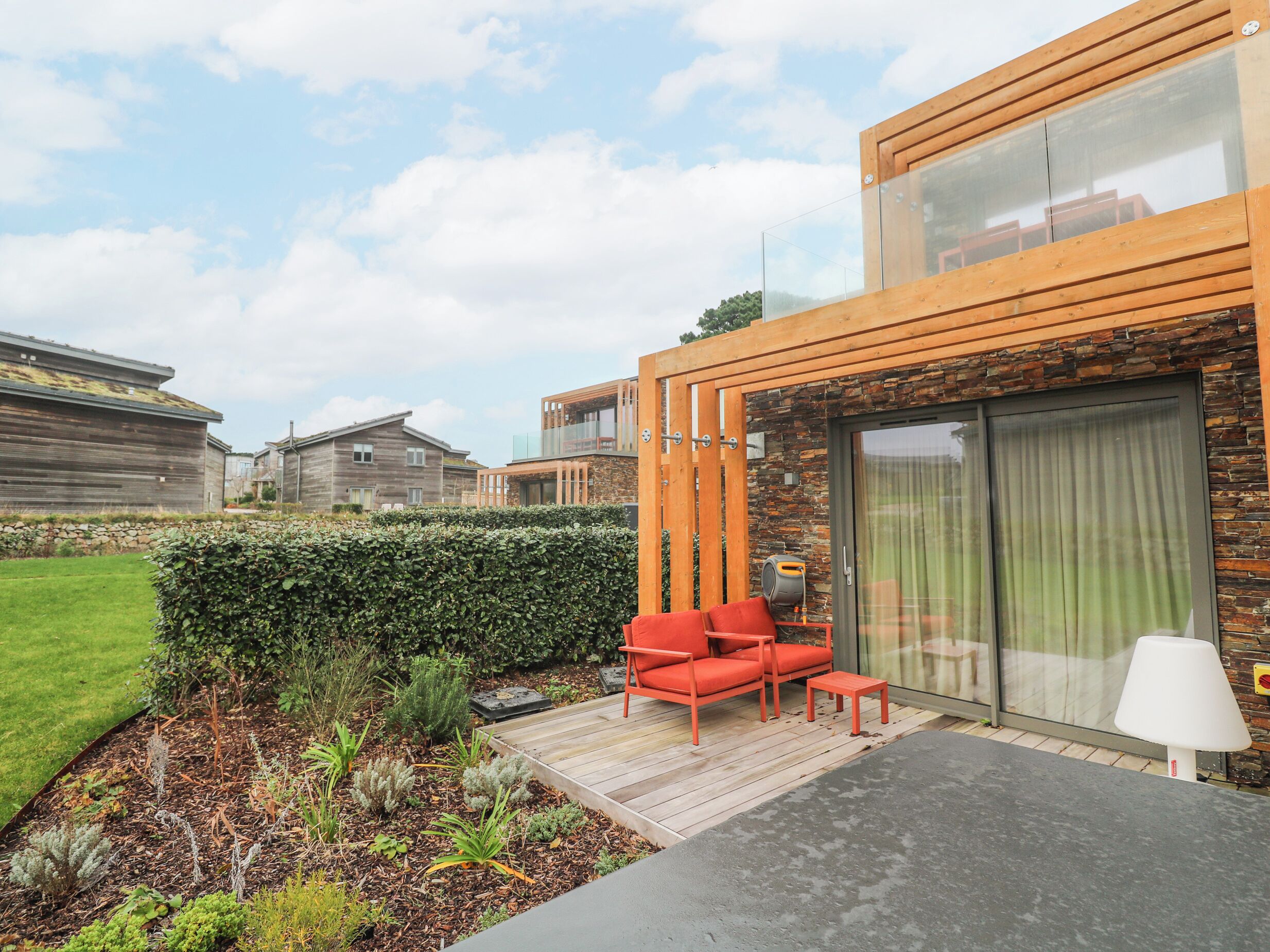 A modern outdoor patio with wooden furniture, a glass door, and a well-maintained garden.