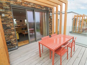 A modern outdoor patio with a wooden deck, a red table and chairs, and a glass sliding door leading to an interior living area with a sofa and a dining table. - Villa 80 (St Ives)
