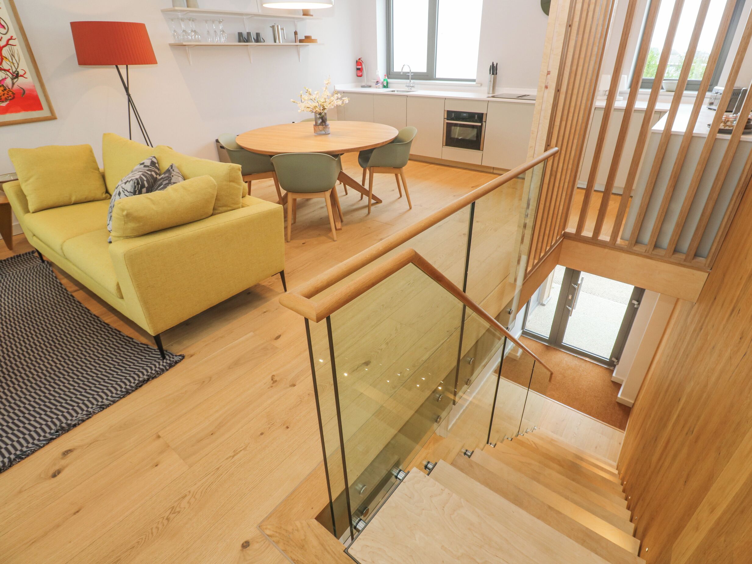 A modern living space with a yellow sofa, a dining table, and a staircase with glass railings.