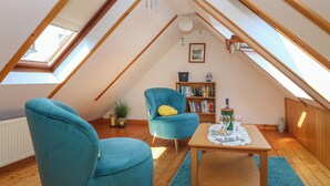 A cozy attic room with a sloped ceiling, wooden beams, a skylight, a blue armchair, a small table with a glass of wine, a bookshelf, and a potted plant. - Jade Cottage (Macduff)