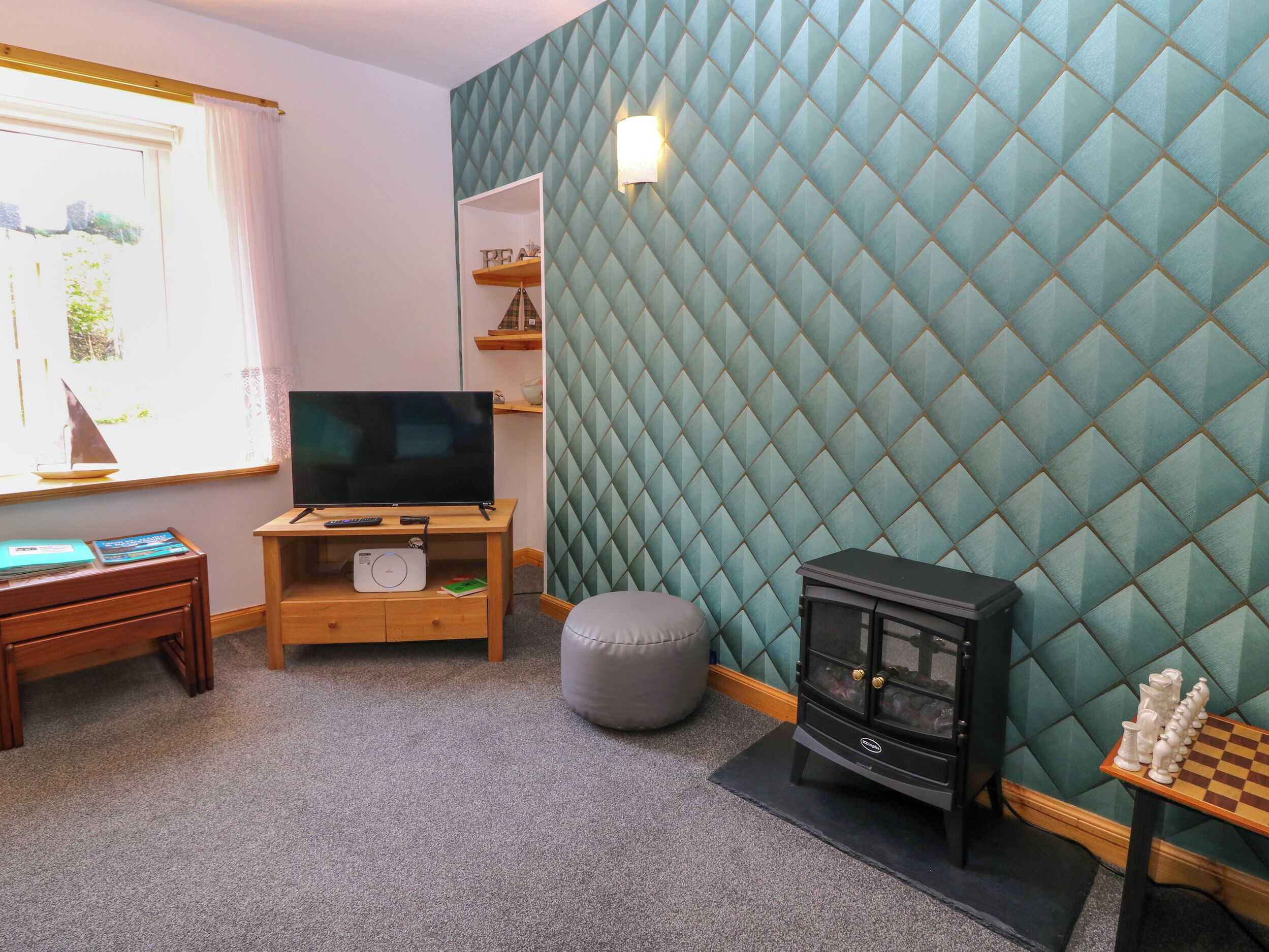 A cozy room with a television on a wooden stand, a small ottoman, a chessboard, and a wall with a diamond-patterned design.