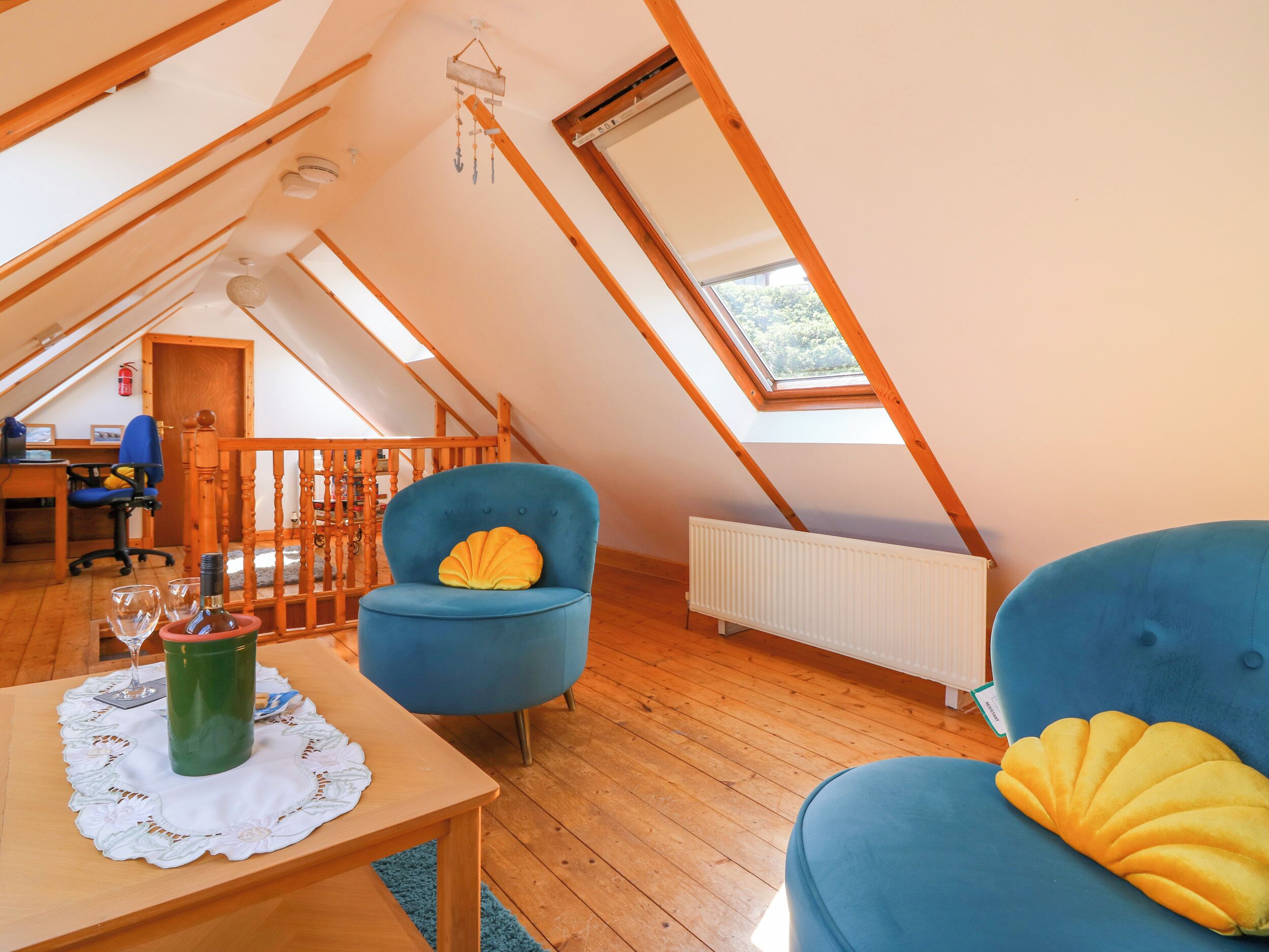 A cozy attic bedroom with a sloped ceiling, wooden beams, a skylight, a blue armchair with a yellow cushion, a wooden table with a glass, and a radiator.