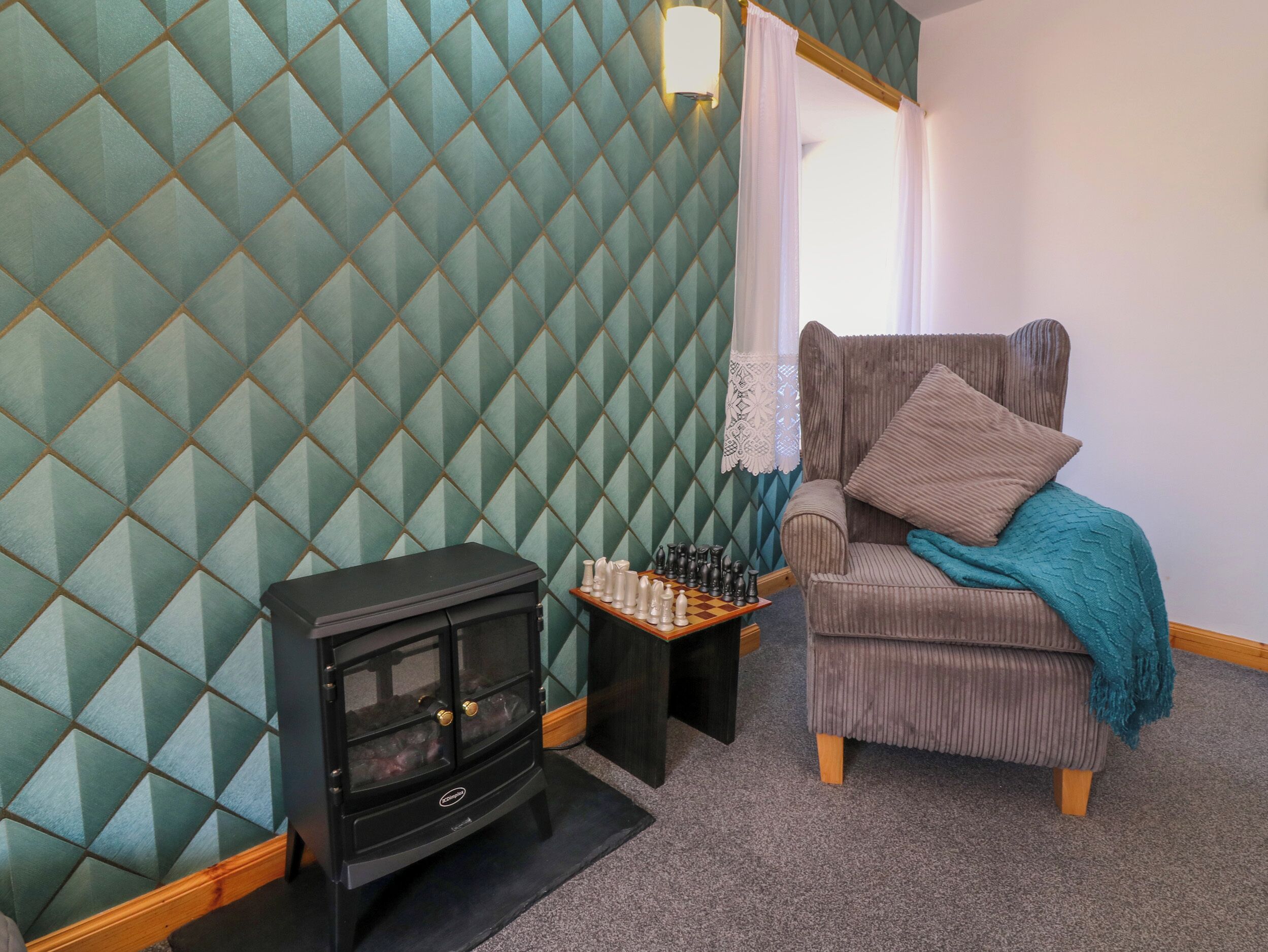 Una habitación acogedora con una pared de diseño verde con motivos de diamantes, una silla de madera con una manta color turquesa y un tablero de ajedrez sobre una mesita.