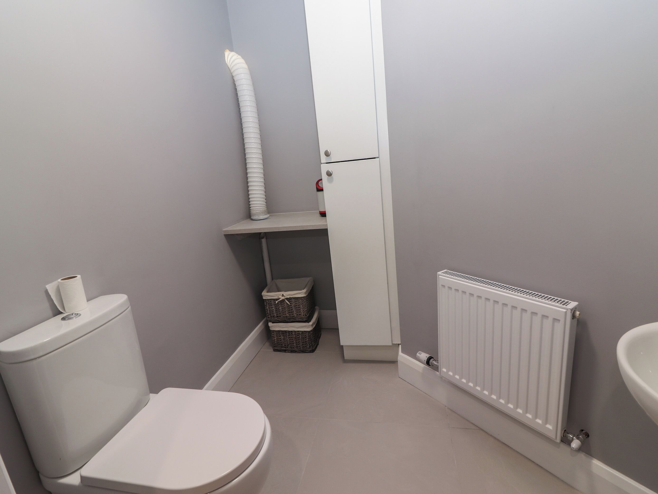 Une petite salle de bain comprenant des toilettes, un radiateur, un lavabo blanc et une étagère avec un panier et un rouleau de papier toilette.