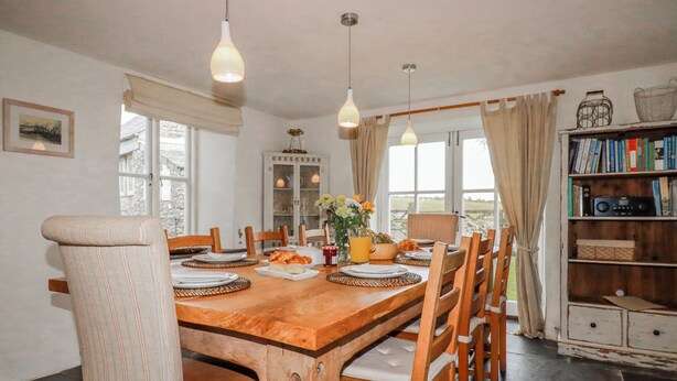 A dining area with a wooden table, chairs, hanging lights, a bookshelf, and a view through the window. - Poldue House (Camelford)