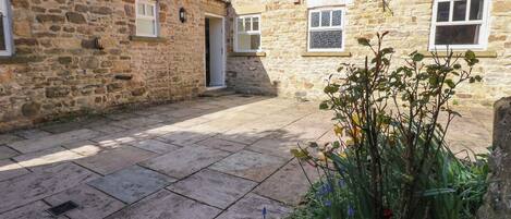 A stone building with a tiled roof, a paved courtyard, and a garden with flowers.
