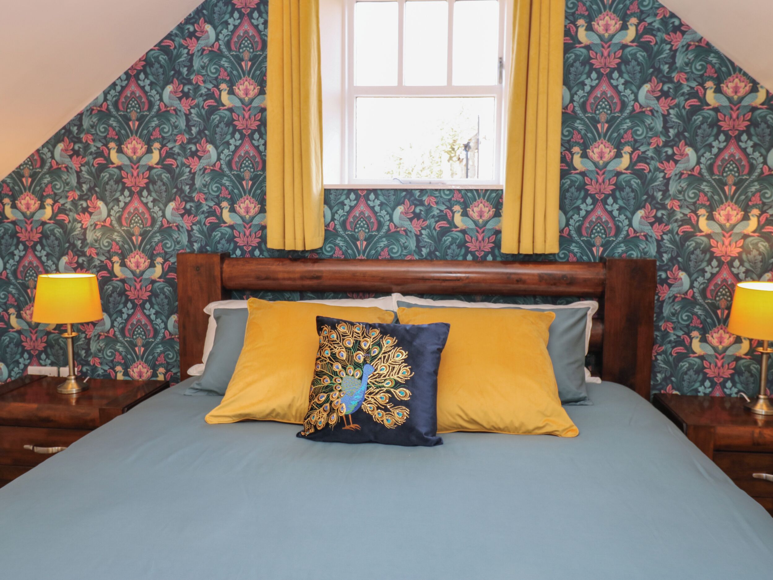 A bedroom with a sloped ceiling, a bed with two pillows, a window with yellow curtains, and patterned wallpaper.