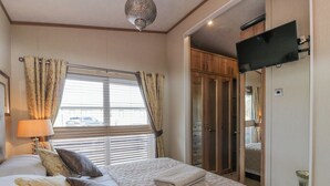 Een slaapkamer met een bed, kussens, een raam met gordijnen, een aan de muur bevestigde televisie en een garderobe.