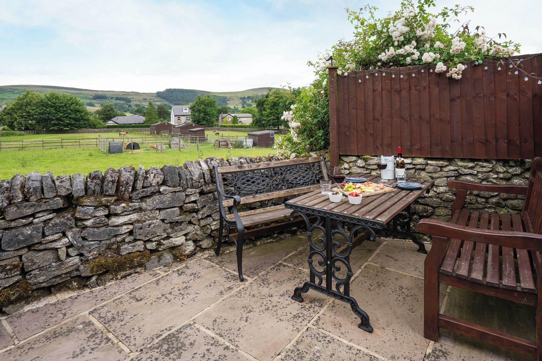 A stone wall patio with a wooden table and benches, a stone wall, and a view of a rural landscape with houses and hills.