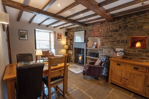 A cozy living room with a stone wall, wooden beams, a fireplace, a bookshelf, and a dining area. - Buttercup Cottage (Hope Valley)