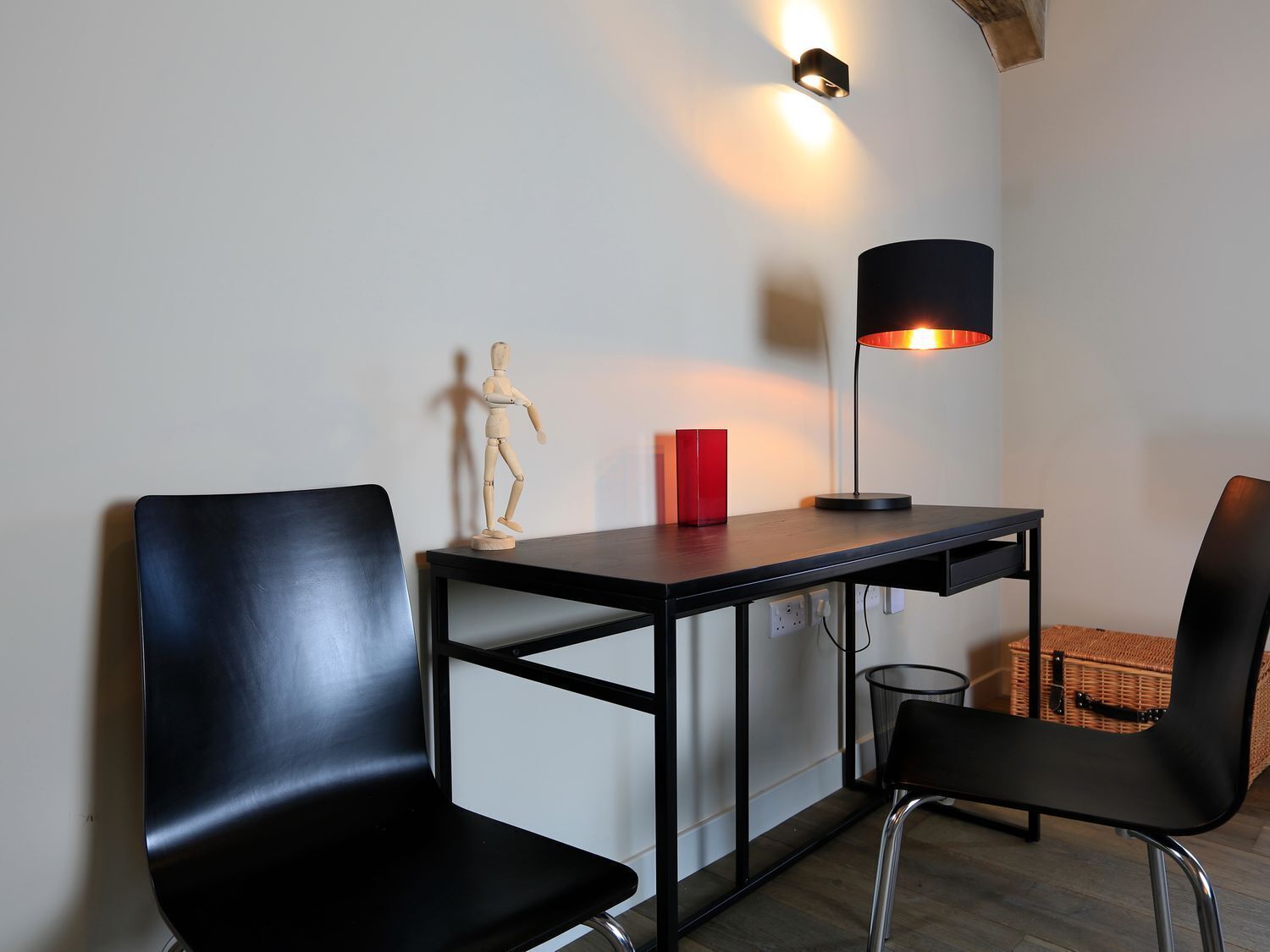A small, modern room with a desk, two black chairs, a red vase, a wooden mannequin, and a floor lamp.